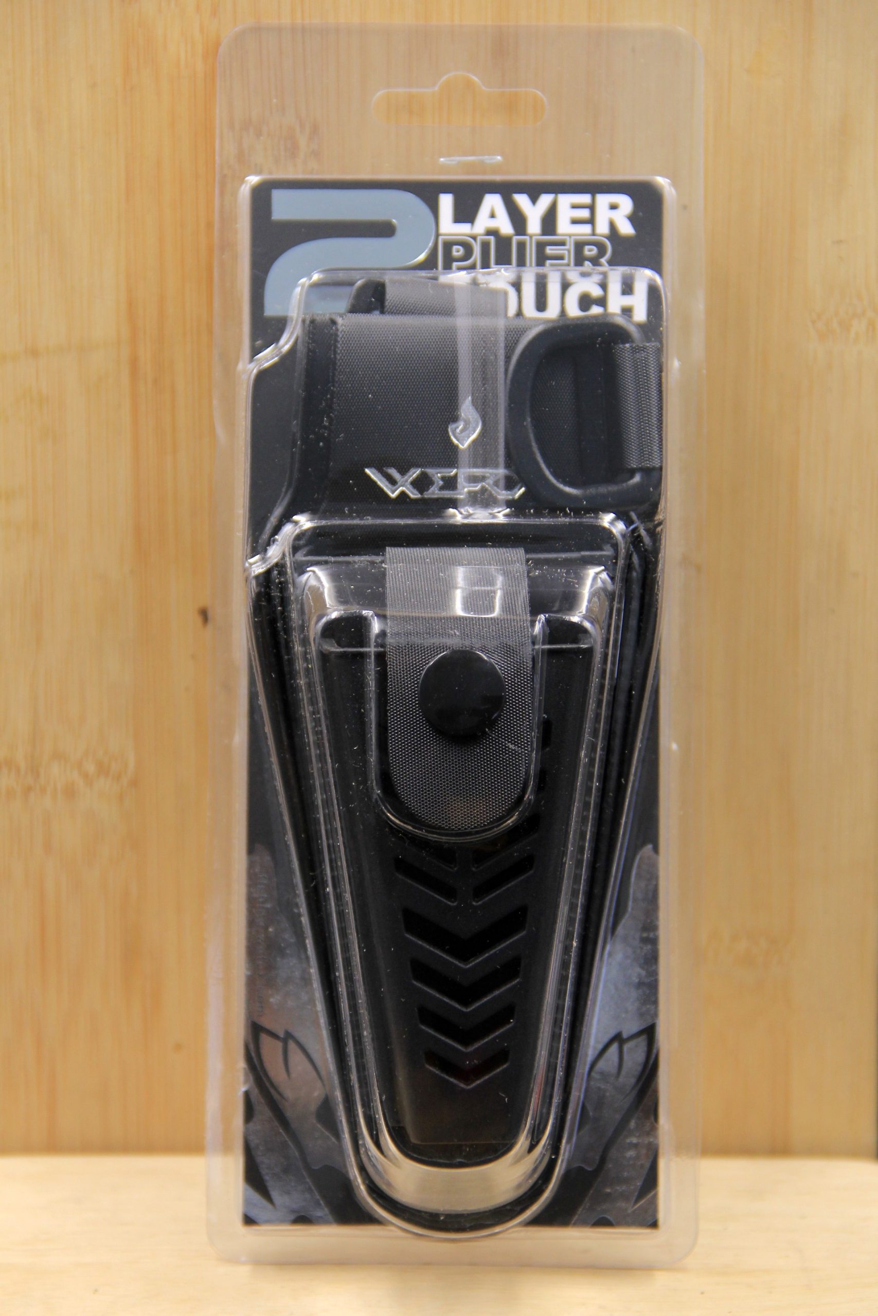 A front-facing product photo of the WEFOX 2-Layer EVA Plier Pouch in black, displayed in its retail clamshell blister packaging, showcasing the textured chevron-patterned EVA front panel, snap-button belt clip closure, nylon webbing upper compartment with Velcro flap, and embossed WEFOX logo, set against a natural bamboo wood background.