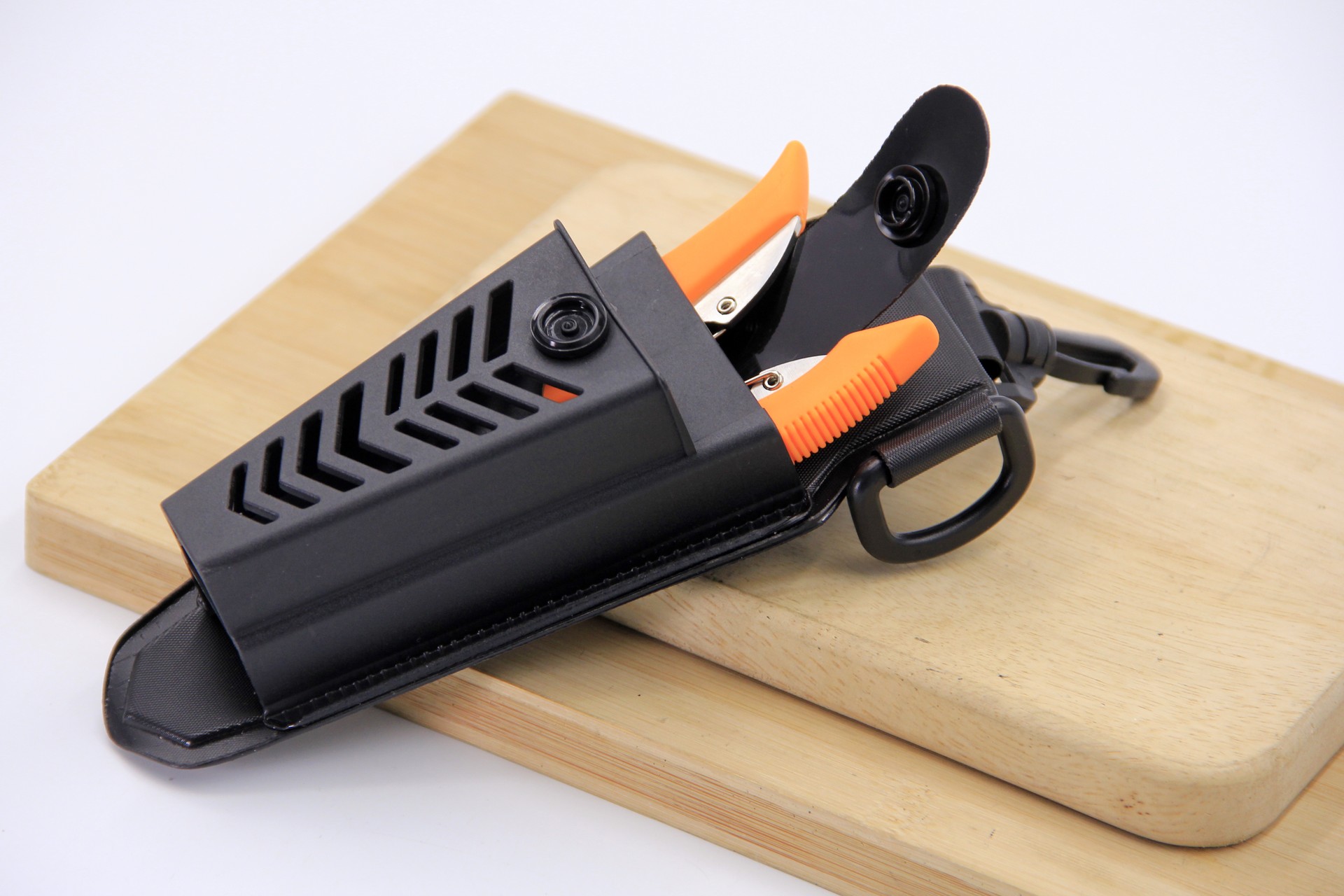A close-up angled view of a WEFOX EVA plier pouch in black with ventilated chevron-pattern cutouts on the front panel, shown resting on a wooden cutting board with a pair of orange-and-black fishing pliers partially inserted and a secondary folding knife stored in the side pocket, alongside an attached black carabiner clip and nylon webbing belt loop.