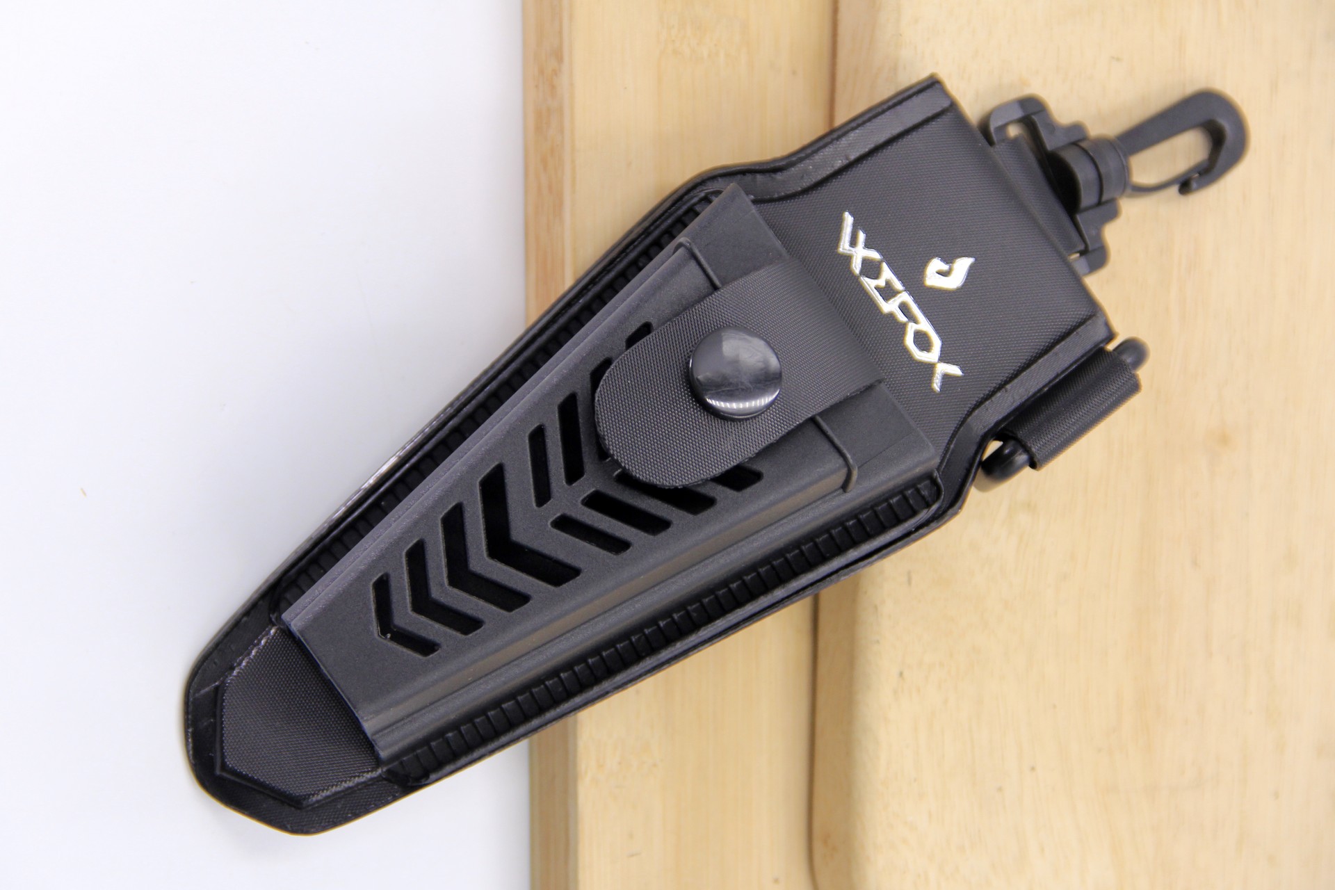 Top-down angled view of a black WEFOX EVA plier pouch featuring a chevron-patterned hard-shell front panel, snap-button retention strap, reinforced nylon edges, and a swivel clip attachment, displayed on a light wooden surface.