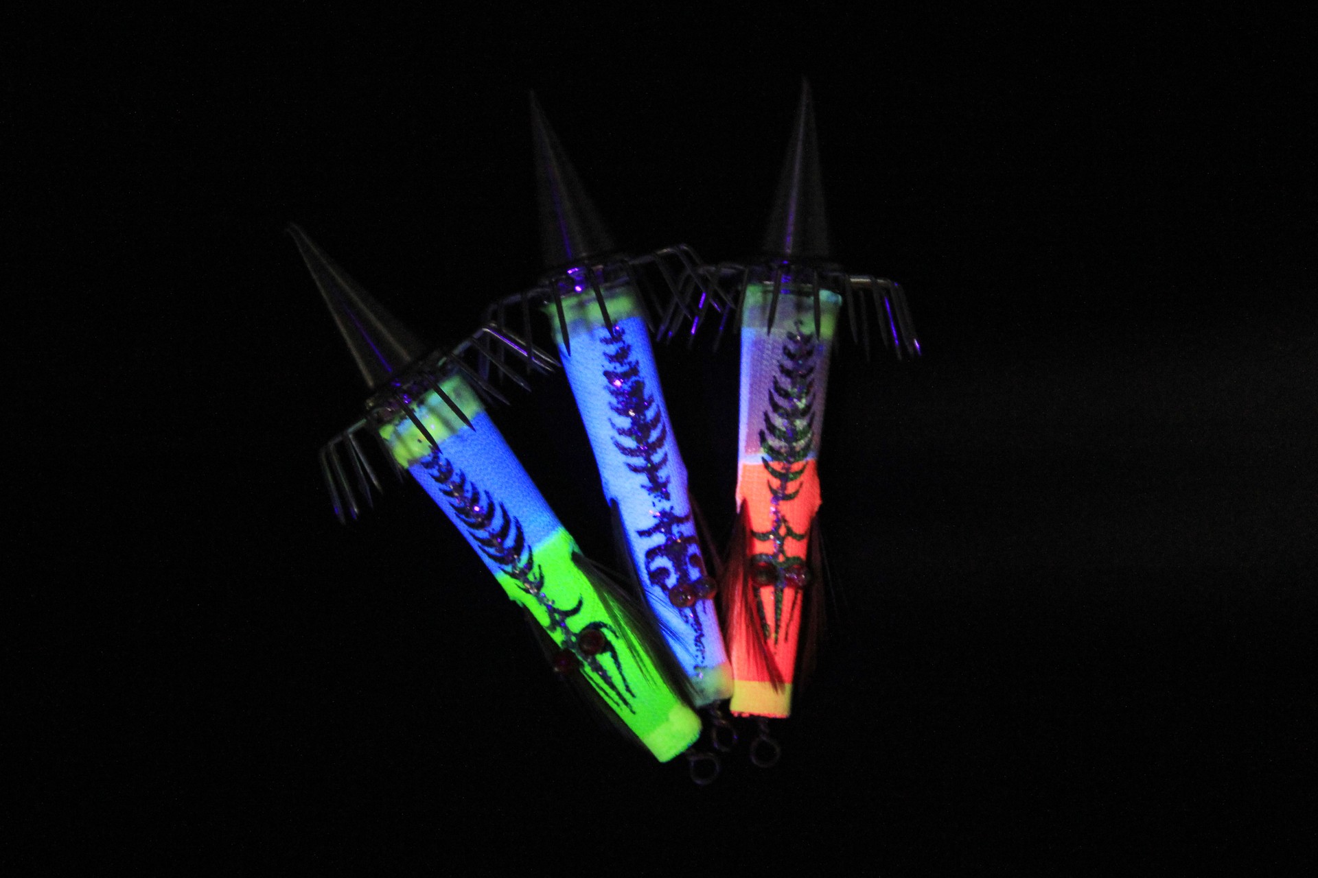 Three glowing Bocean Candat Twist Squid Jig Bottom Sinkers in green, blue, and red with luminous cone bodies and umbrella hooks on black background.